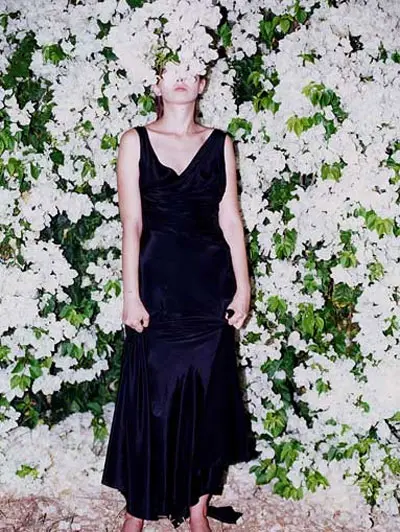 שמלה שחורה עם מחשוף וי, נשענת על קיר מכוסה בפרחי בוגנויליה לבנים מתוך הקטלוג האחרון של כתומנטה