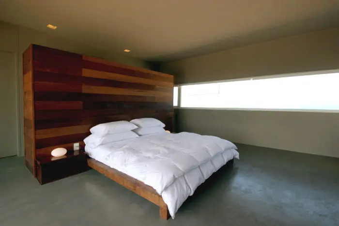 חדר שינה מינימליסטי עם חלון רחב לצפיה בנוף. מאחורי המיטה קיר עץ שמצידו האחורי משמש כאזור אחסנה