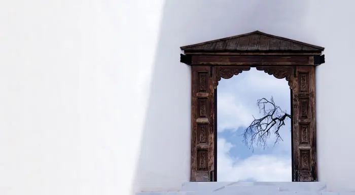 ענף עץ זית בן 200 שנים מציץ מבעד לשער כניסה עשוי מעץ הנמצא בין שתי החומות, דרכו יורדים אל הבית שנבנה במדרון וכוסה באדמה