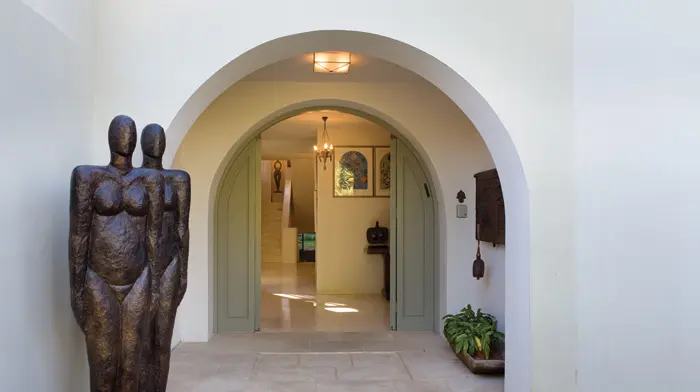 שני פסלים מעשיה ידיה של גילה, הנראים זהים, עשויים מחומרים שונים, ניצבים על מפתן הבית. ברקע דלתות הכניסה המקומרות וגרם המדרגות