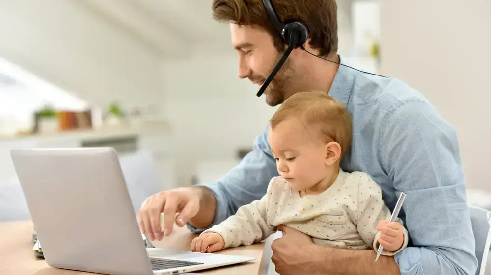 גבר יושב מול לפטופ עם אוזניה ותינוקת על הברכיים