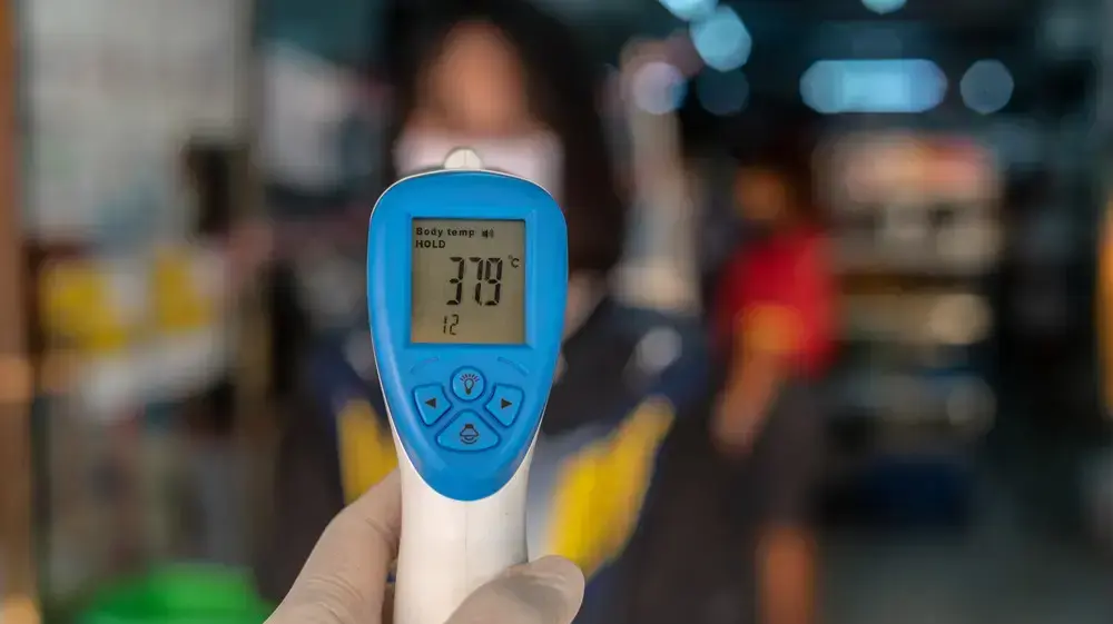 יד אוחזת באקדח מדחום שעל הצג שלו מופיעה המדידה 37.9 מעלות צלזיוס