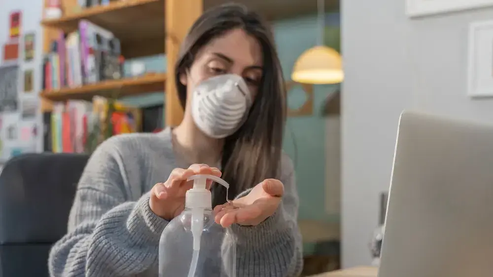 אישה יושבת מול לפטופ עם מסיכה ומחטאת ידיים