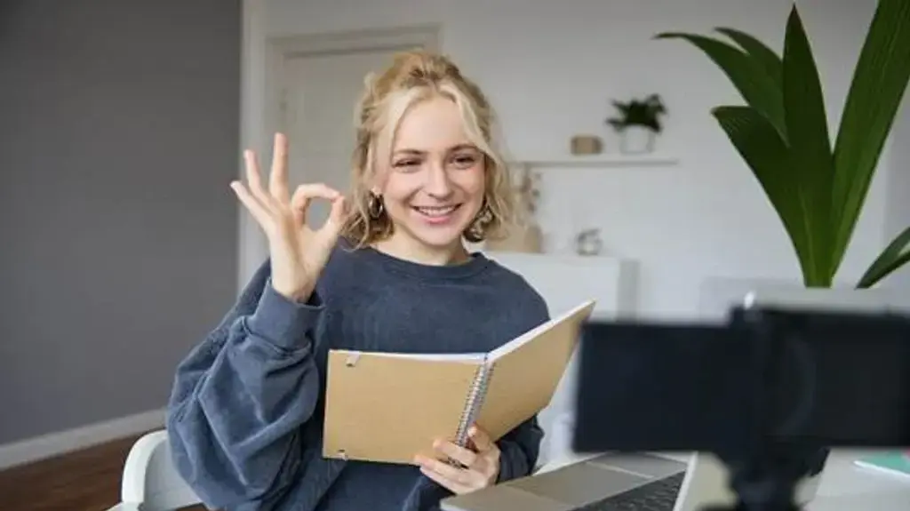 אישה מחזיקה ספר ומביטה בחיוך למחשב, עם מחווה של שביעות רצון עם האצבעות